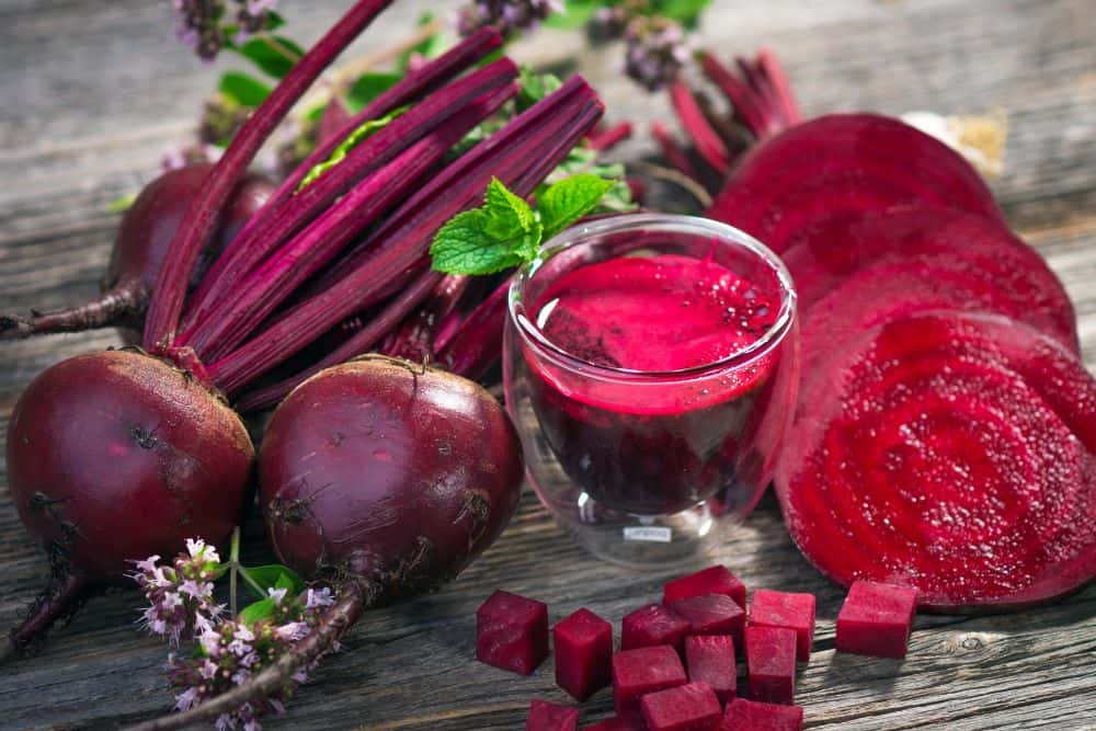Ein Glas Rote Beete Saft neben Rote Bete Knollen auf einem Holztisch