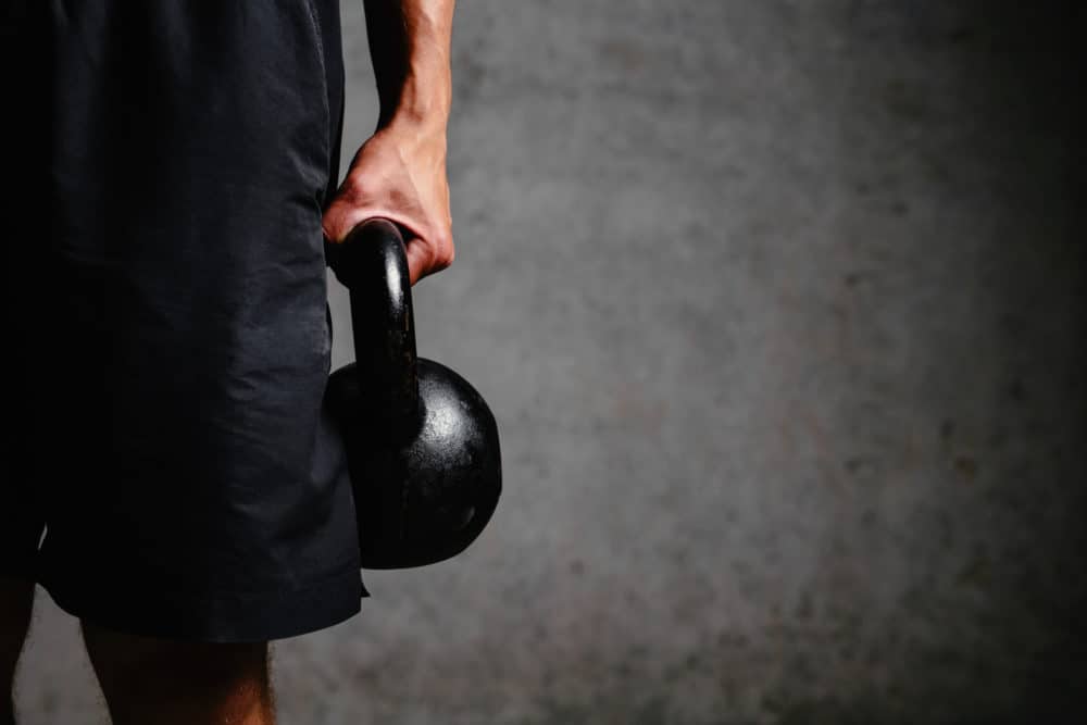 Athlet mit schwarzer Hose in einer Hand eine Kettlebell (Kugelhantel) vor schwarzer Wand