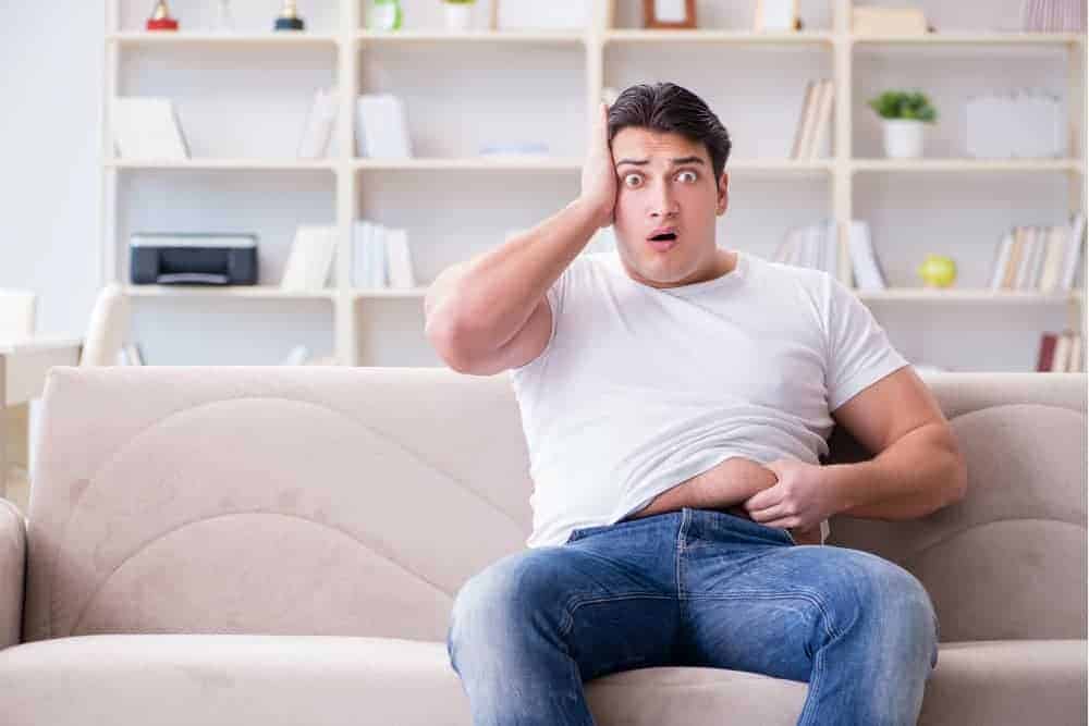 Bauchfett loswerden junger Mann mit Übergewicht sitzt auf dem Sofa und greift sich ans Bauchfett