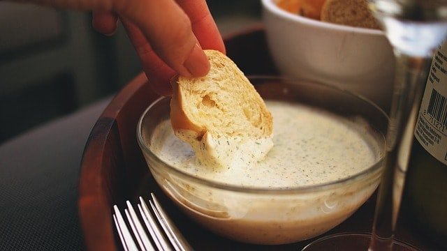 Aioli in weißer Glasschale mit Weißbrotstück, das von einer Frauenhand eingetunkt wird