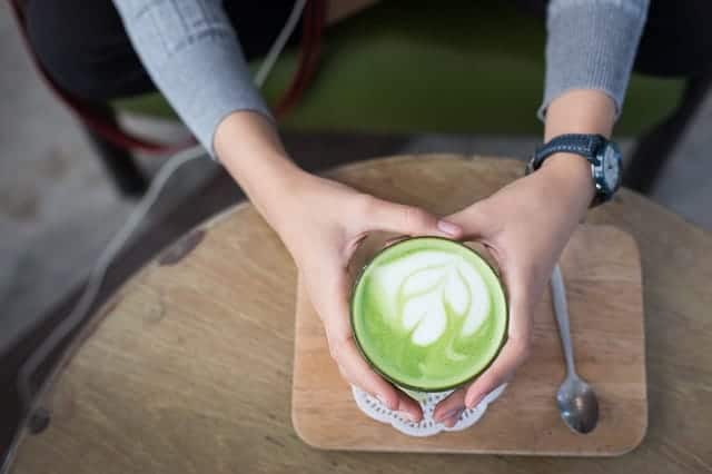 Frauenhände umfassen Matcha-Latte im Hintergrund Holzbrett und Holztisch